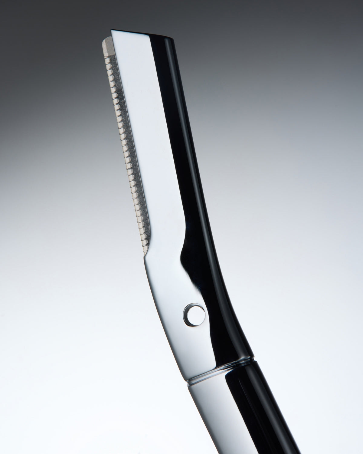 A close-up studio shot of a professional eyebrow razor with a polished silver metallic blade and a glossy black handle, set against a smooth gray gradient background.