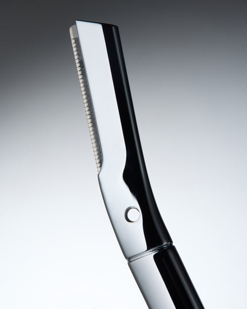 Bild 2 in Galerieansicht laden. A close-up studio shot of a professional eyebrow razor with a polished silver metallic blade and a glossy black handle, set against a smooth gray gradient background.
